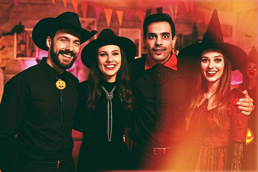Four adults at a Halloween party posing together. One man wears an all-black cowboy outfit with a jack-o’-lantern bolo tie, while a woman beside him wears a black outfit with a sparkling bolo tie featuring a black cat.